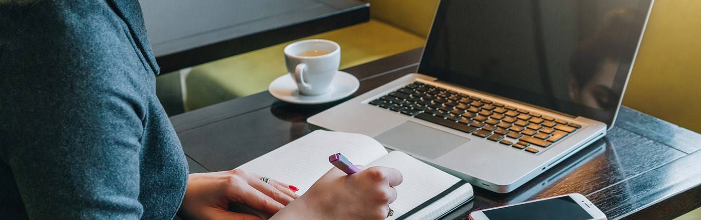 person writing down on her notebook with laptop in front