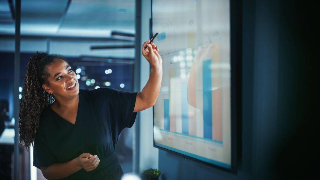 Company Operations Director Holds Sales Meeting Presentation for Employees and Executives. Creative Black Female Uses TV Screen with Growth Analysis; Charts; Ad Revenue. Work in Business Office.